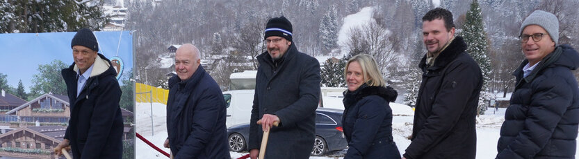 Eine Gruppe von sechs Personen beim Spatenstich vor einem verschneiten Hintergrund und einem Bauplan des Resorts.