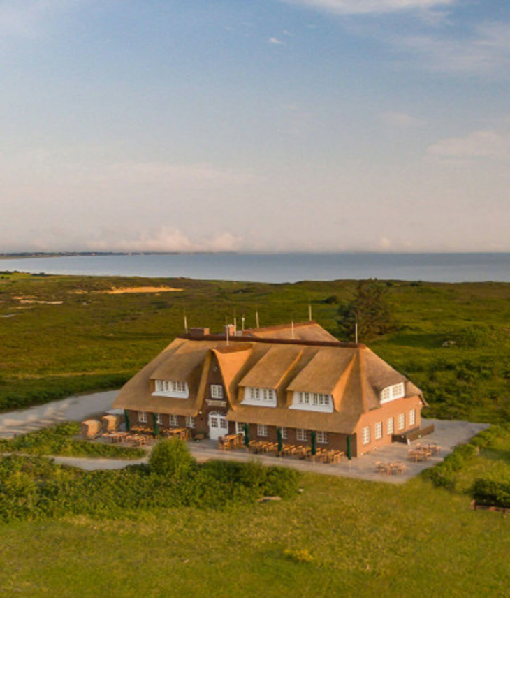 Reetgedecktes Hotel Landhaus Severin*s auf Sylt am Meer, umgeben von grüner Landschaft und blauem Himmel.