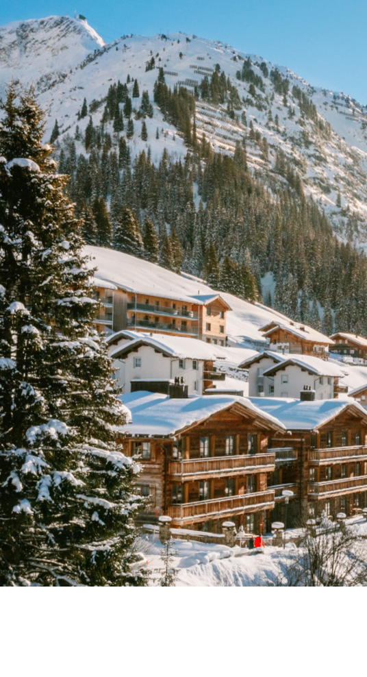 Verschneite Alpenlandschaft mit Sonne über Hotel Severin*s in Lech, umgeben von Bergen und Bäumen.