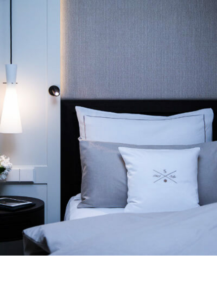 Modernes Hotelzimmer mit elegantem Bett, grauen und weißen Kissen, stilvoller Beleuchtung und Blumen auf dem Nachttisch.