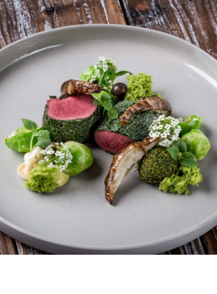 Gourmetgericht mit rosa gebratenem Fleisch, grünen Kräutern und Gemüse, kunstvoll auf einem grauen Teller angerichtet.