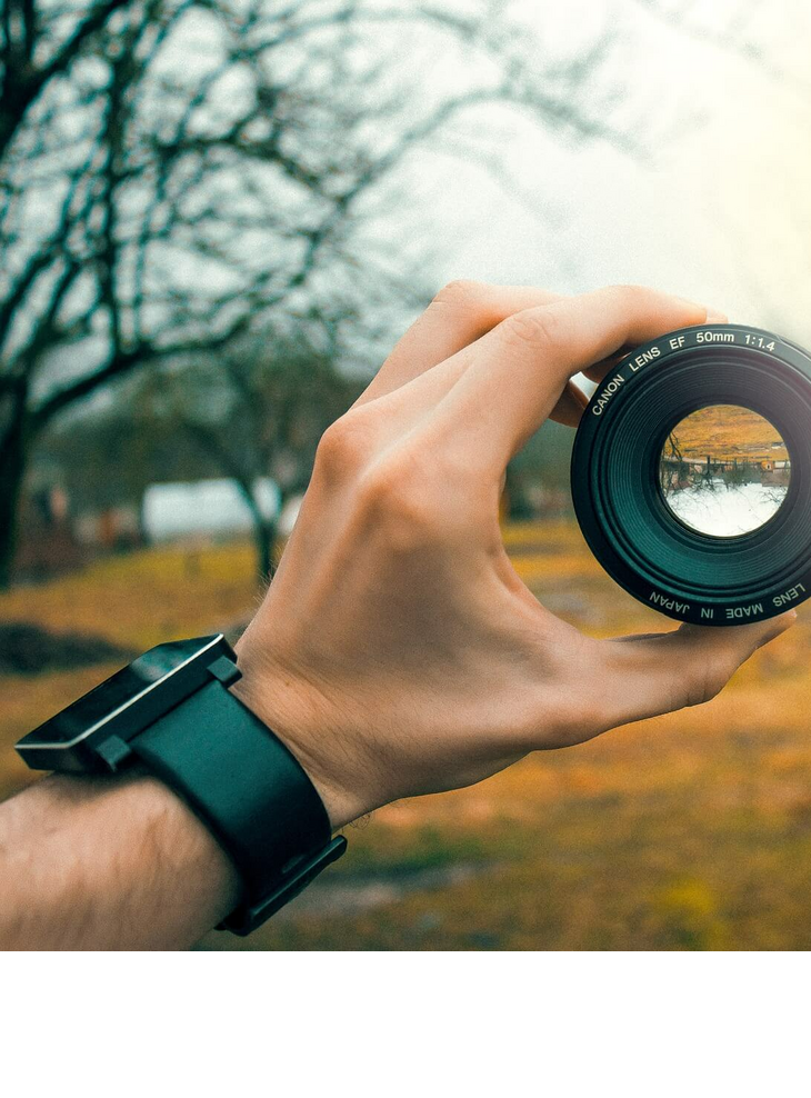Hand hält Kameraobjektiv, das Landschaft fokussiert; unscharfer Hintergrund mit Bäumen.